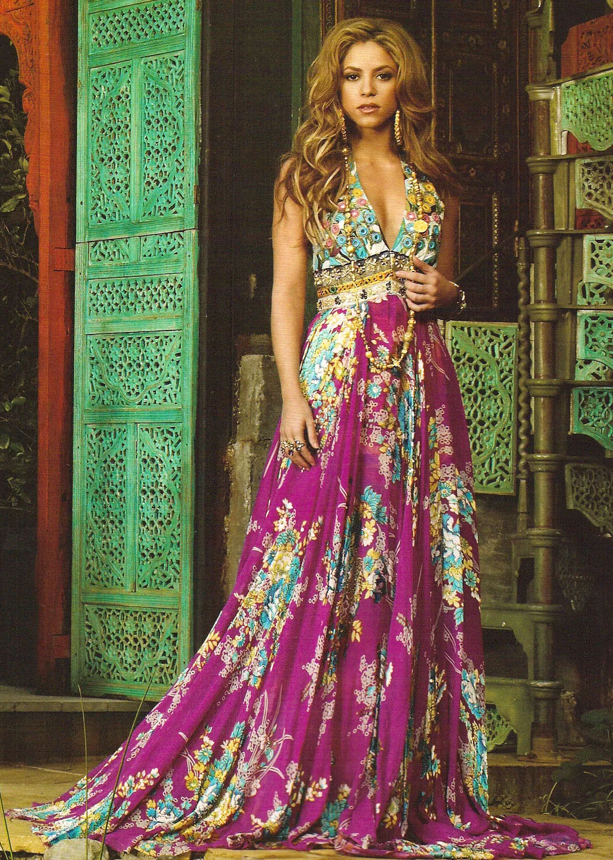 A woman wears a floor-length magenta floral halter gown, standing before carved green wooden doors.